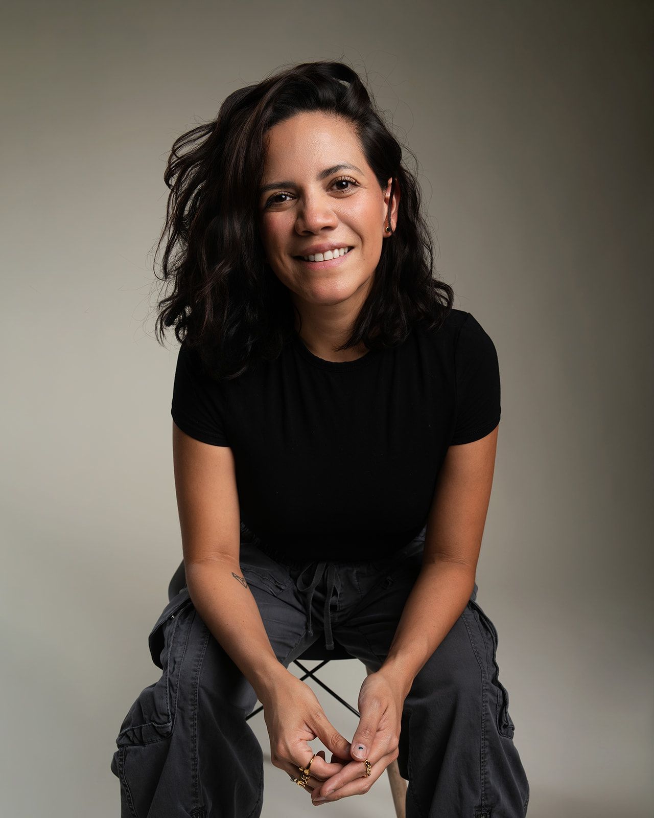 Woman with dark curly hair smiling warmly while seated, wearing a black t-shirt and cargo pants.