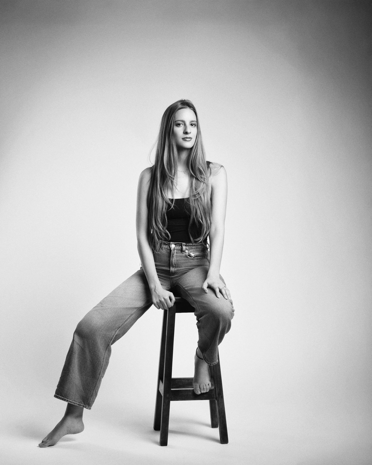 Black and white studio portrait of a young woman with long hair seated barefoot on a wooden stool.