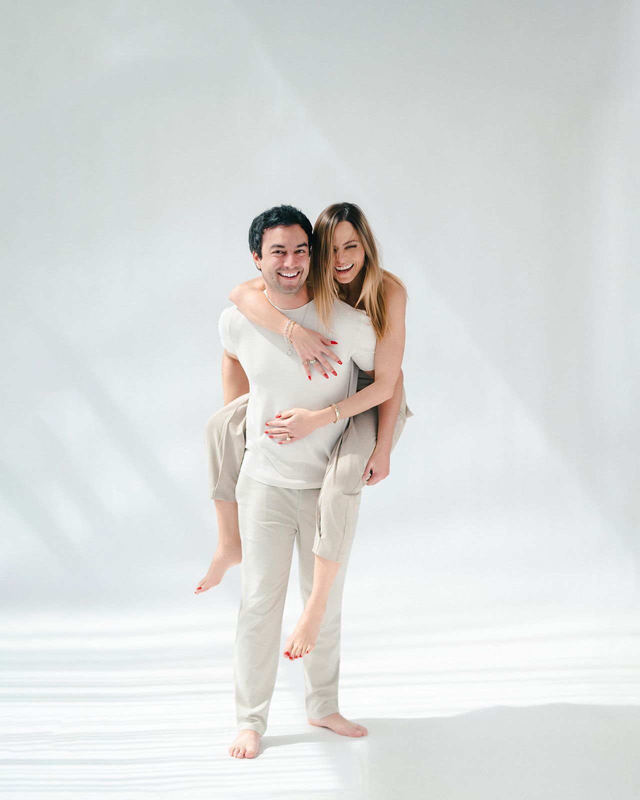 A smiling couple in white outfits, man giving woman a piggyback ride against a white background.