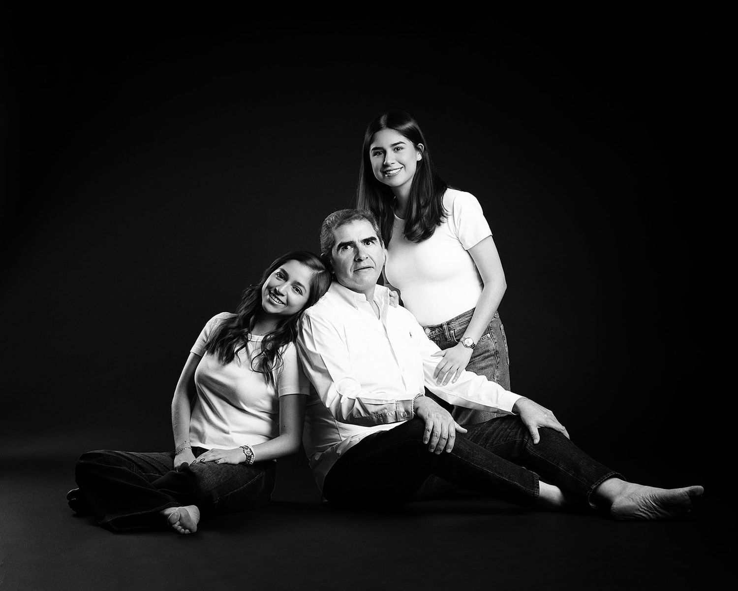 Black and white studio portrait of father seated on floor with two daughters leaning on him.