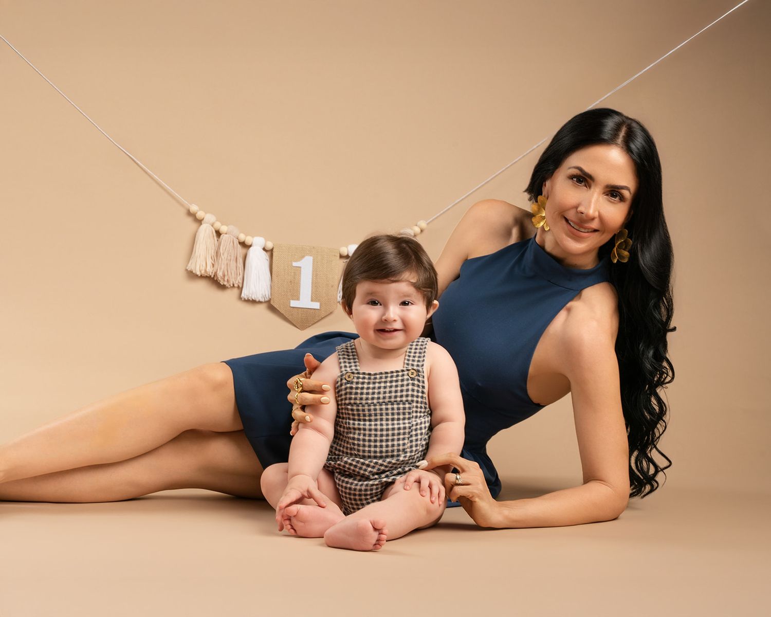 A mother in a blue dress poses with her smiling baby at a first birthday cake smash photo session.