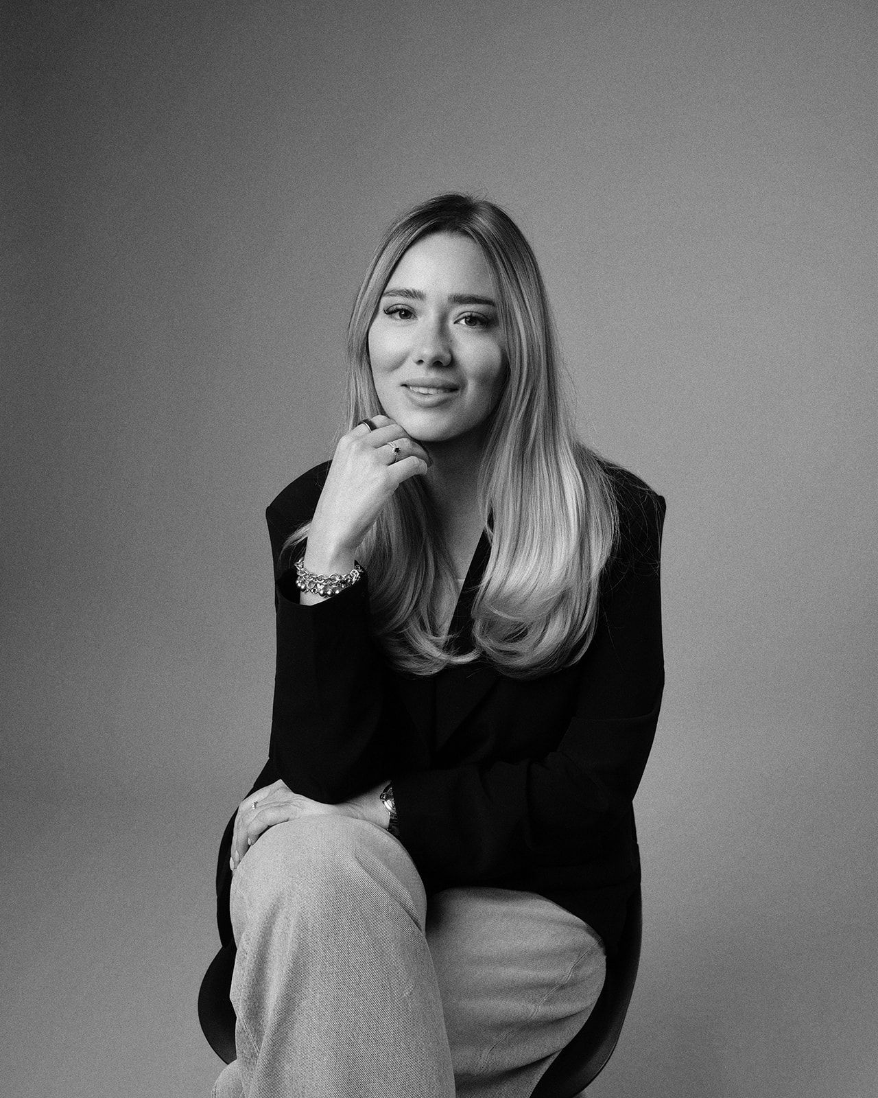 Black and white portrait of a blonde woman in a black blazer, seated and posing elegantly in a studio setting.