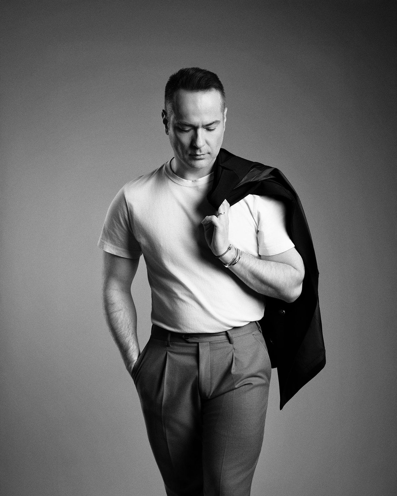 Black and white studio portrait of a man in a white t-shirt holding a blazer over his shoulder.