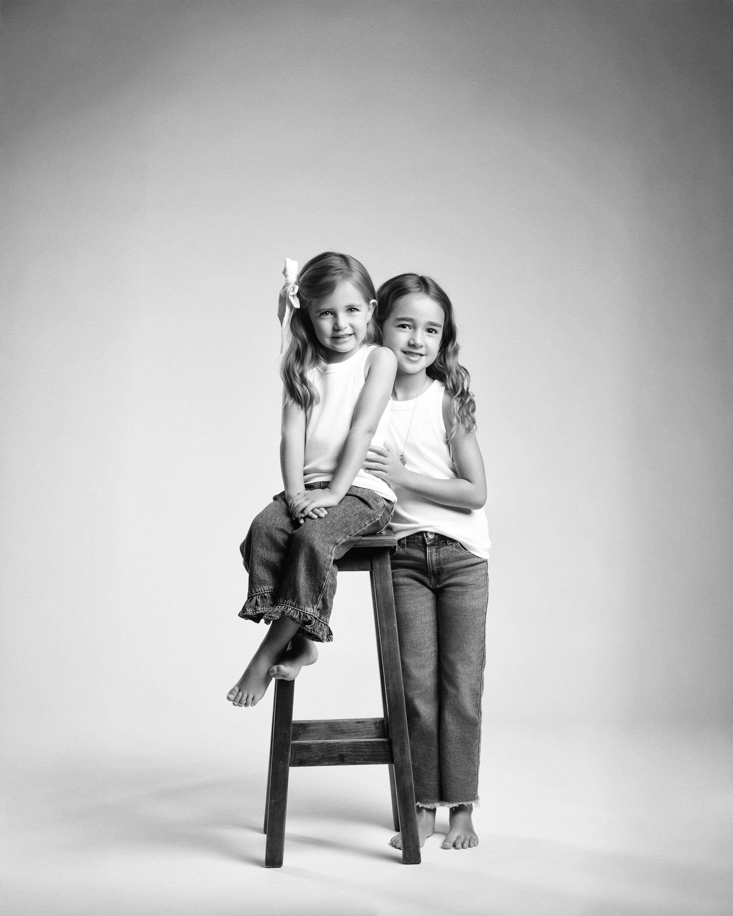 Black and white studio portrait of two young girls, one sitting on a stool and one standing beside her.