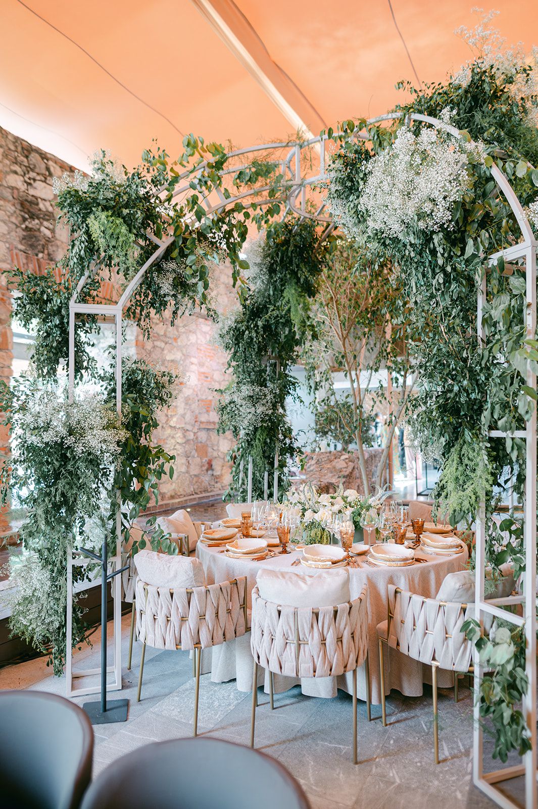Lush greenery wedding reception arch frames elegant round table with blush linens and gold accents.
