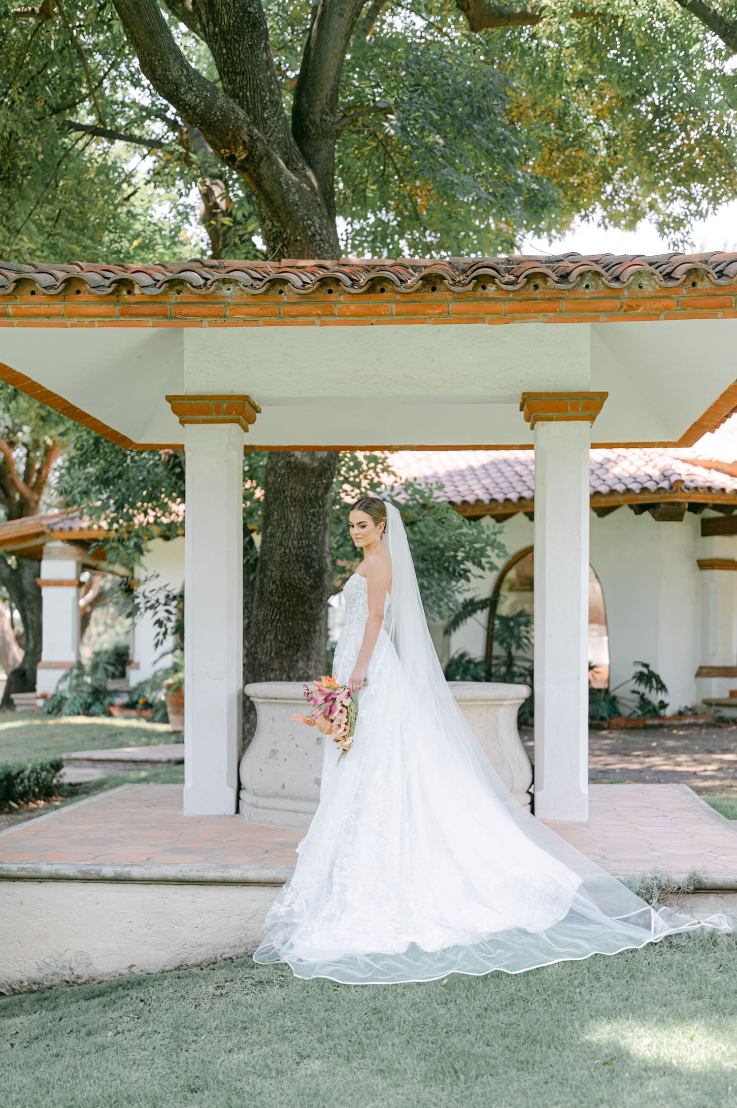 Bride in elegant lace gown with long veil posing under Spanish-style pavilion at wedding venue.