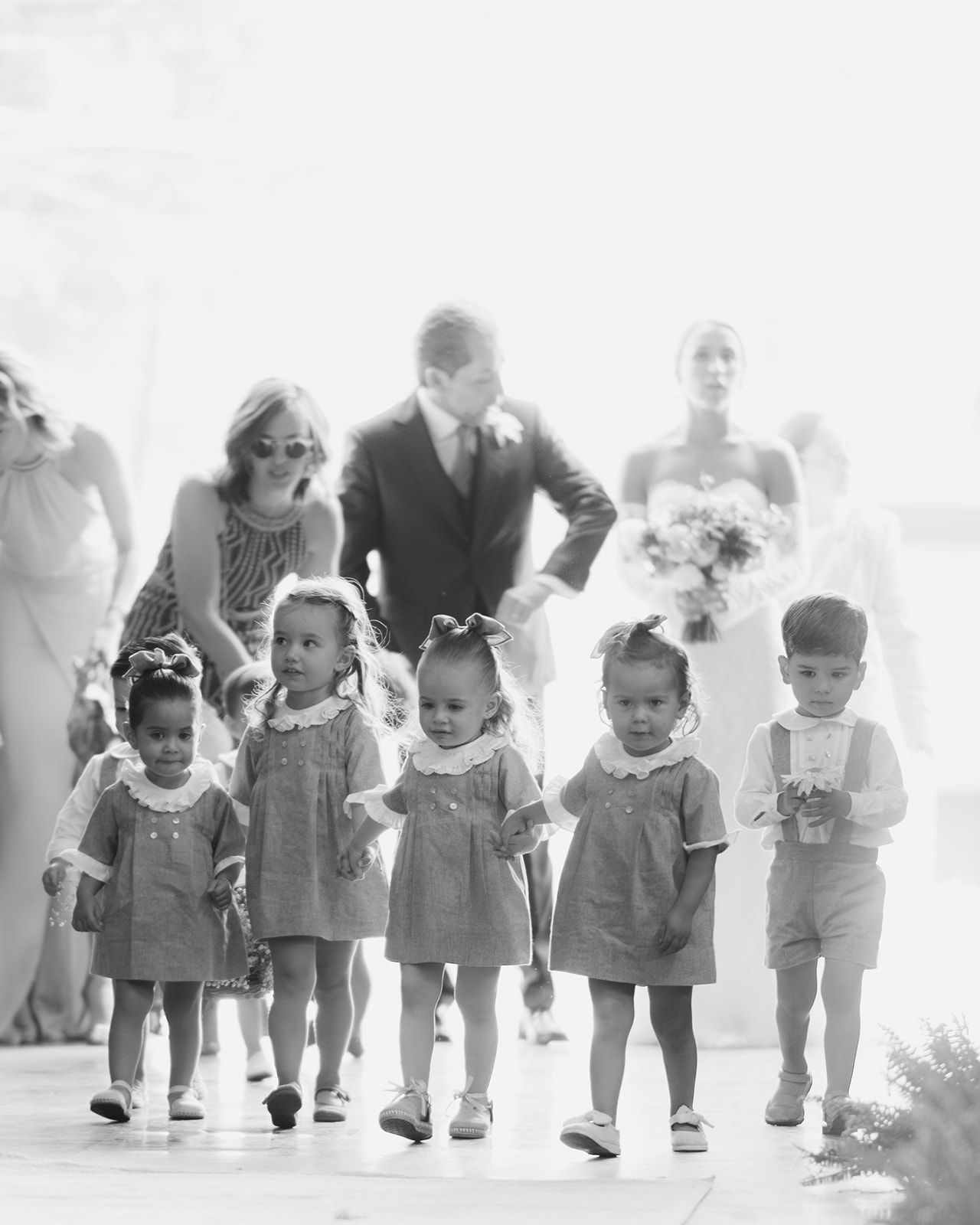 Black and white photo of young flower girls and ring bearers walking down wedding ceremony aisle.