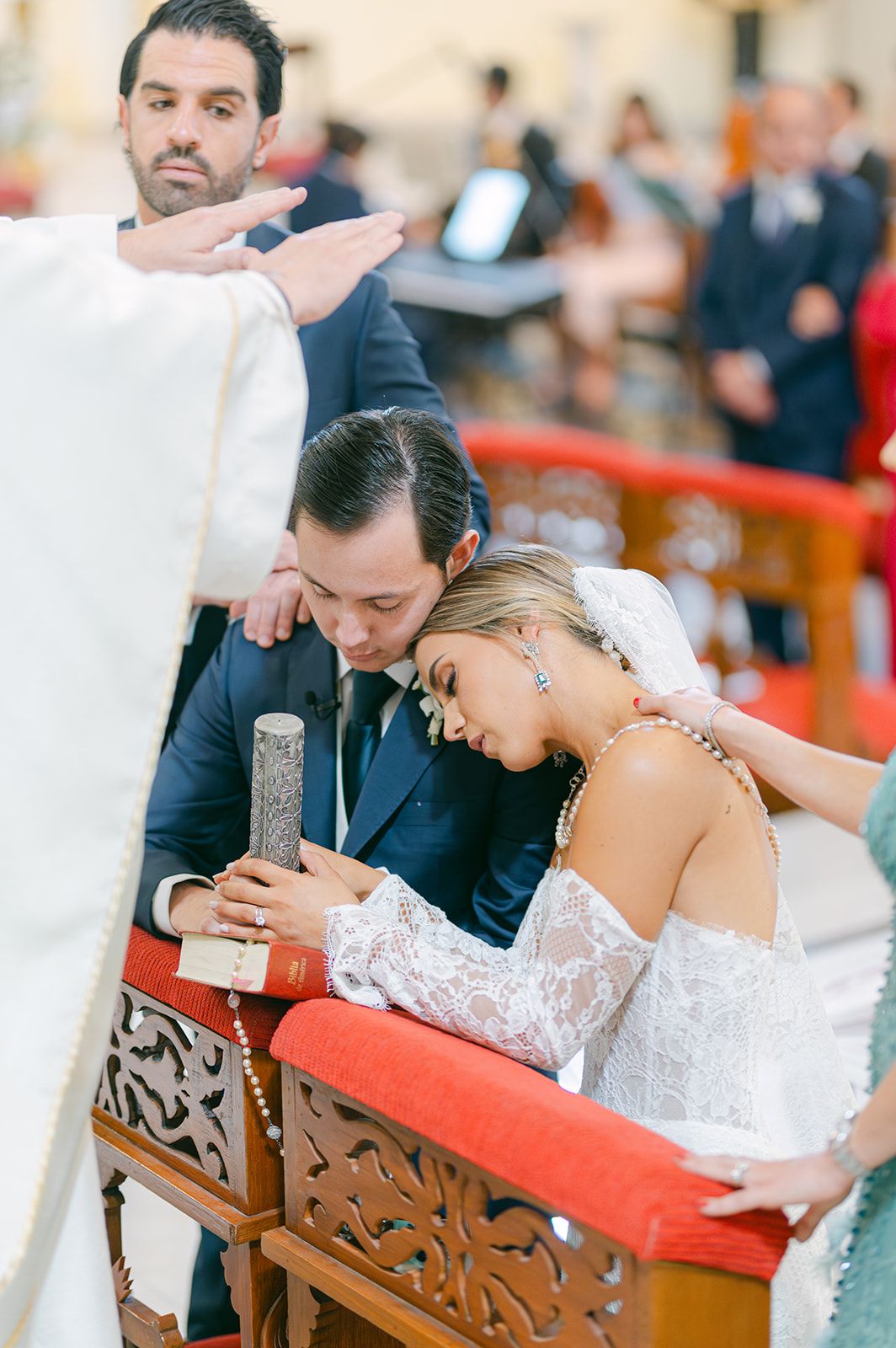 Bride rests head on groom's shoulder as priest blesses them during wedding ceremony.