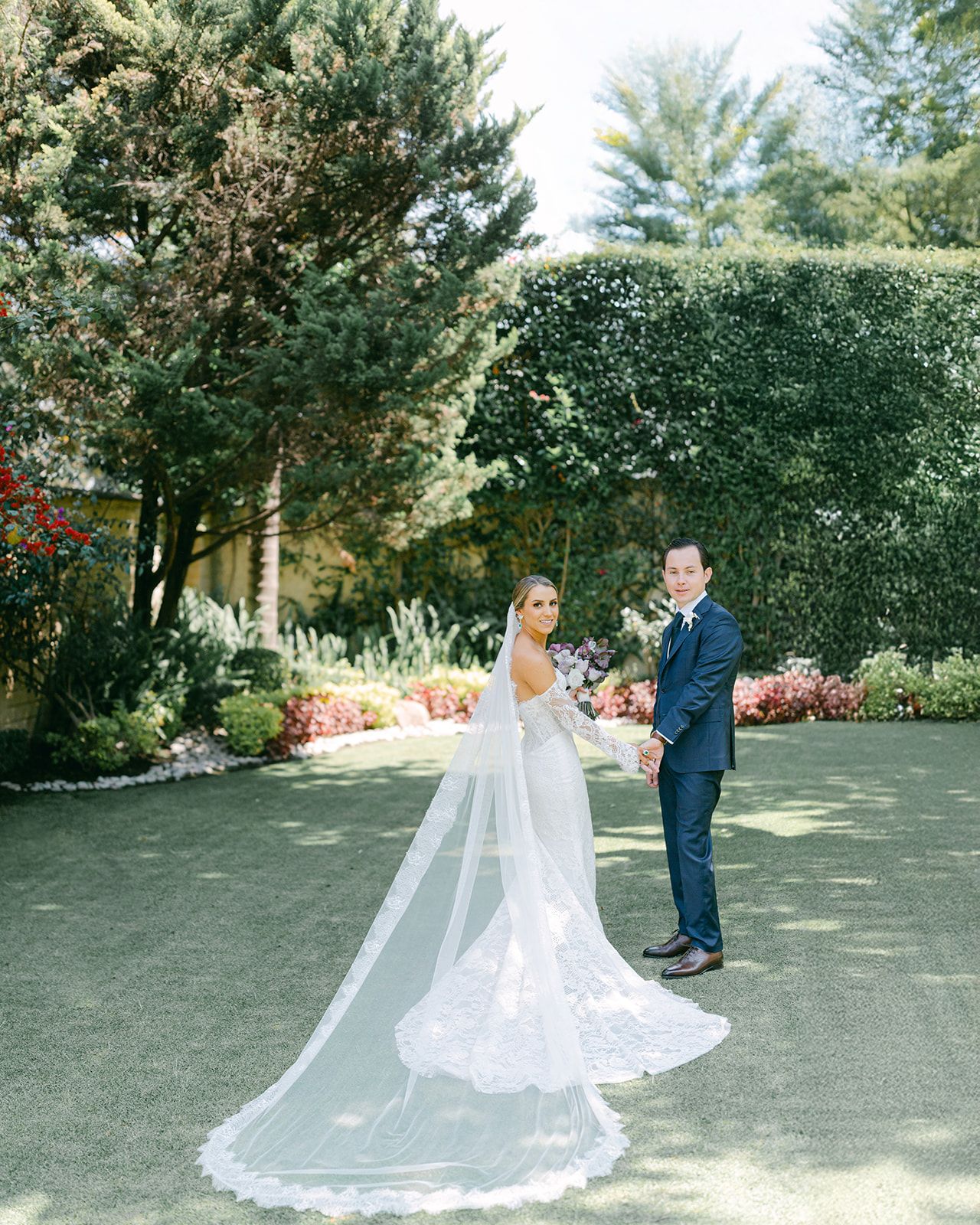 Bride and groom posing in formal garden, bride in lace gown with long cathedral veil trailing.
