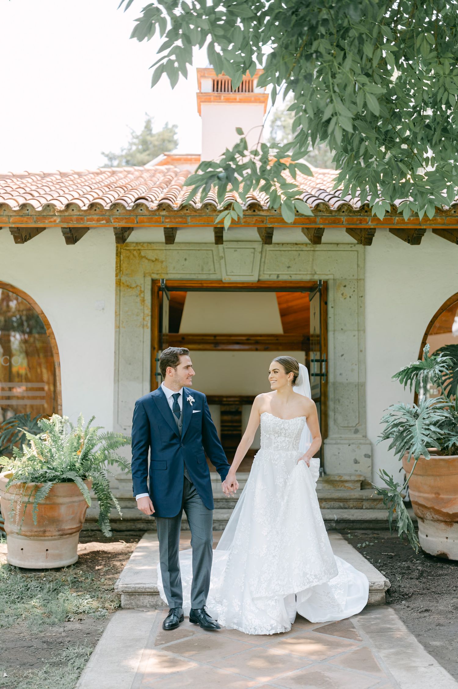 Bride and groom holding hands walking outside Spanish colonial wedding venue with terracotta roof.