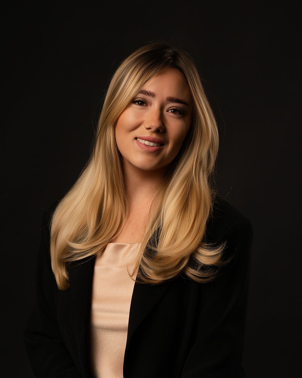 Professional headshot of young blonde woman in black blazer against dark studio background.