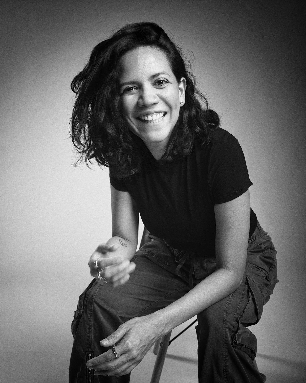 Black and white close-up of a joyful dark-haired woman seated, wearing a black t-shirt.