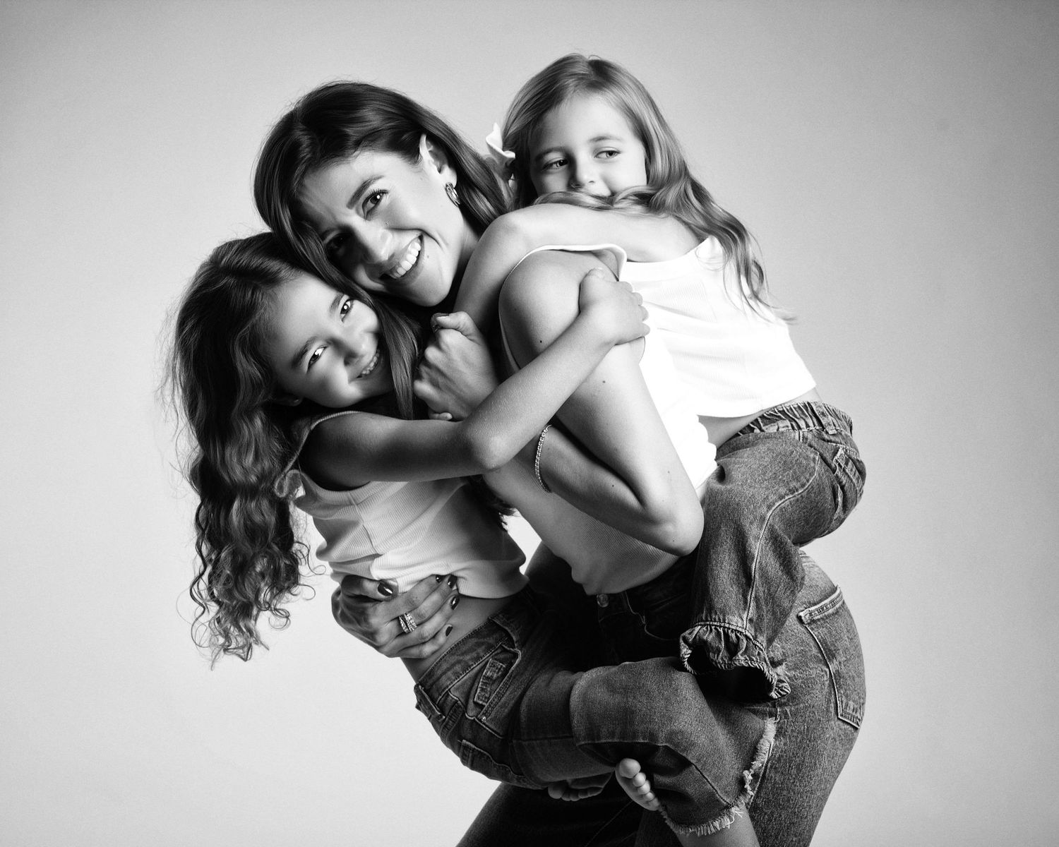 Black and white studio photo of a mother hugging her two young daughters playfully.