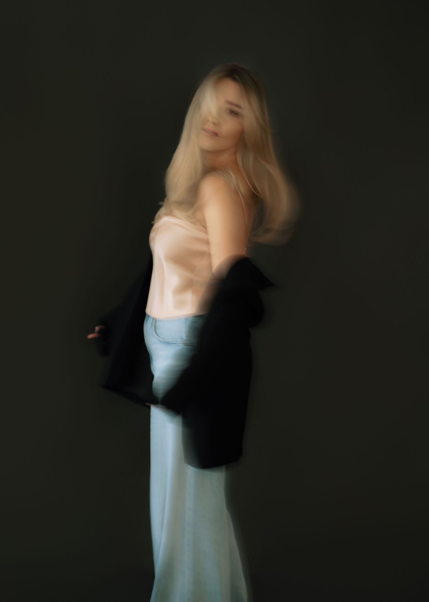 Blurred artistic photo of a blonde woman in satin top and jeans holding a black blazer.
