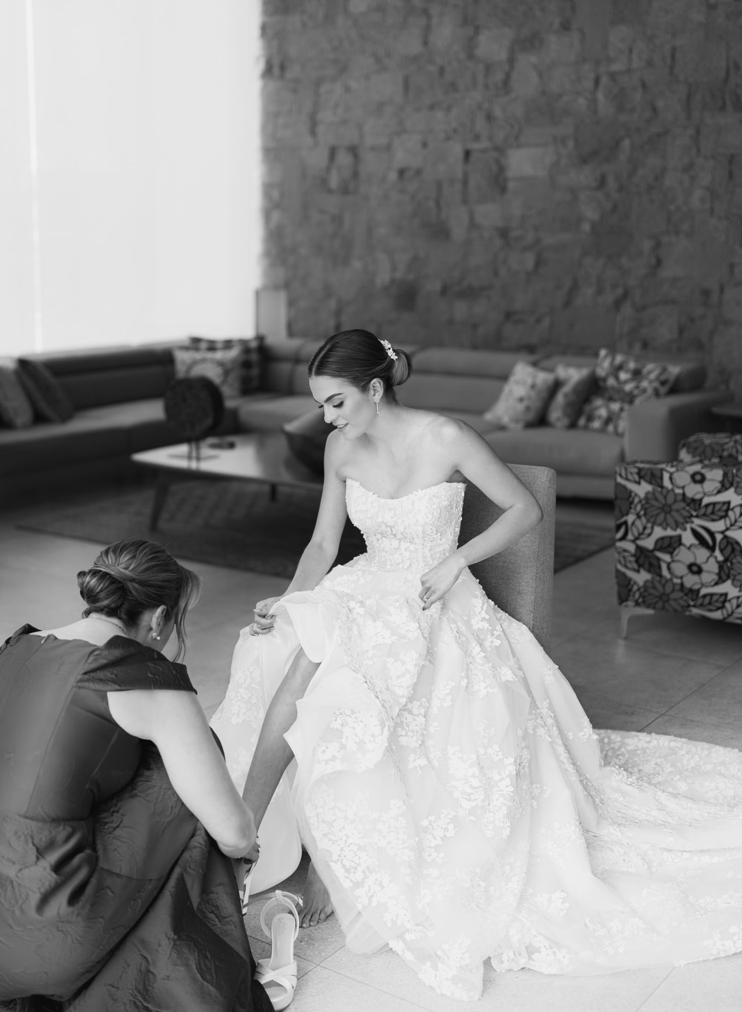 Black and white photo of a bridesmaid helping a bride in a strapless lace gown with her shoes.