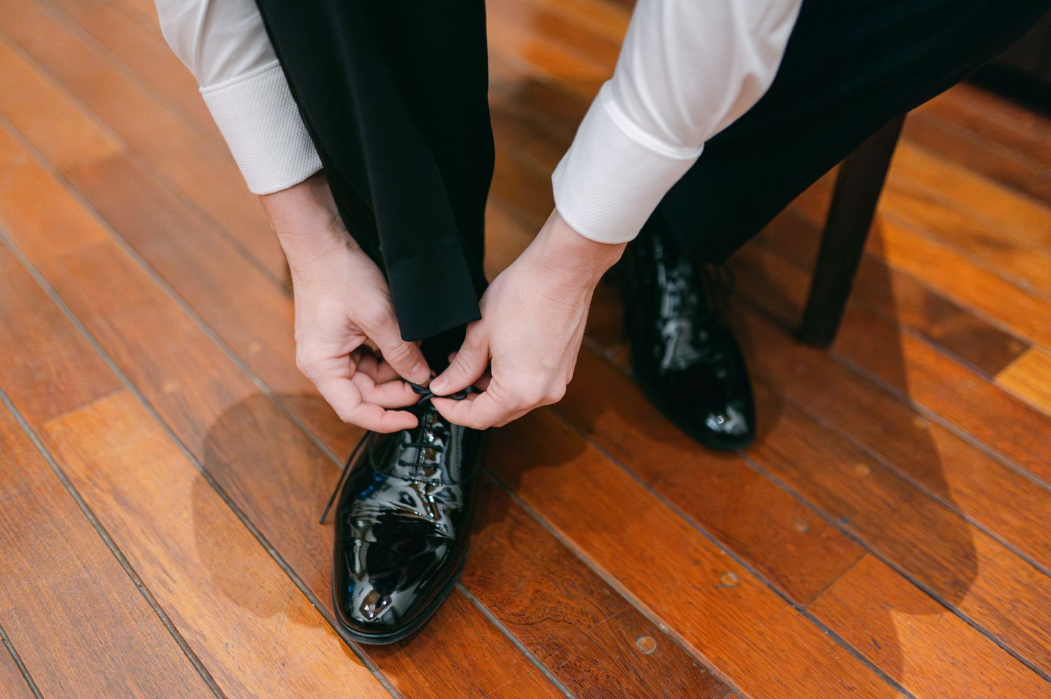 Groom tying black patent leather dress shoes on hardwood floor before wedding.