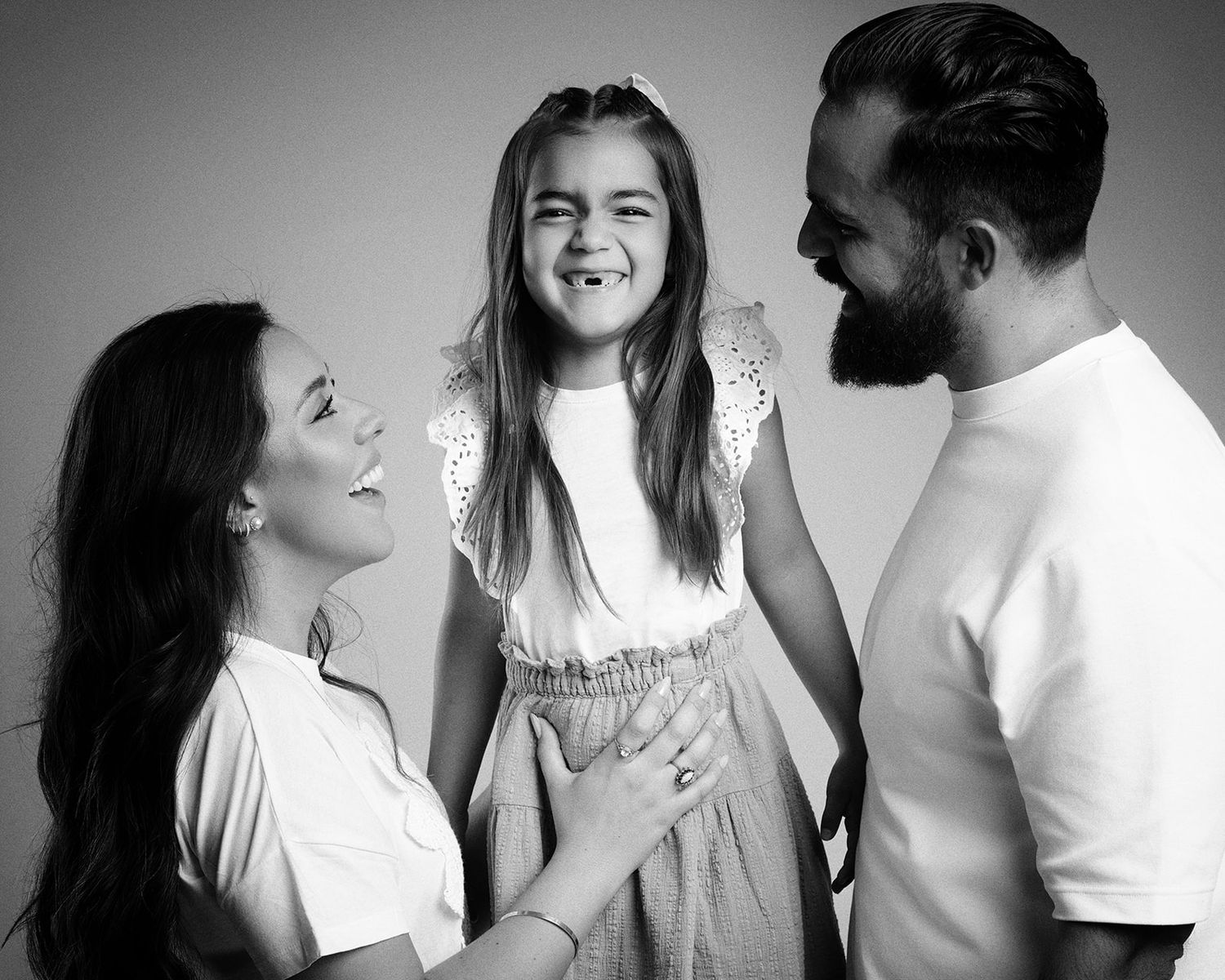 Black and white portrait of a mother and bearded father smiling at their laughing young daughter in a studio.