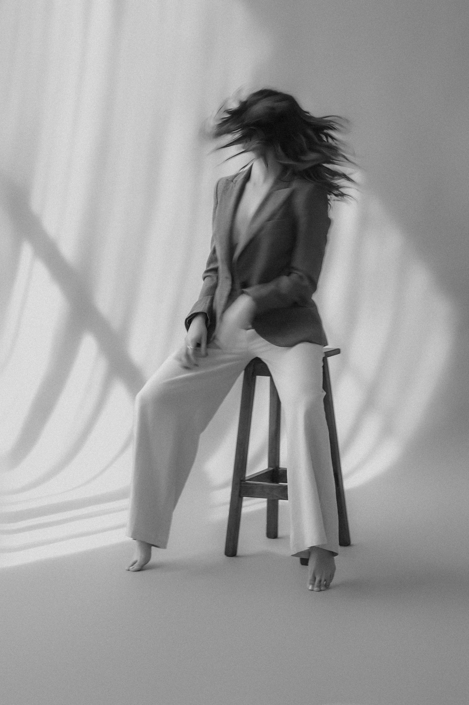 Black and white photo of woman in blazer and wide-leg pants on stool with hair in motion.