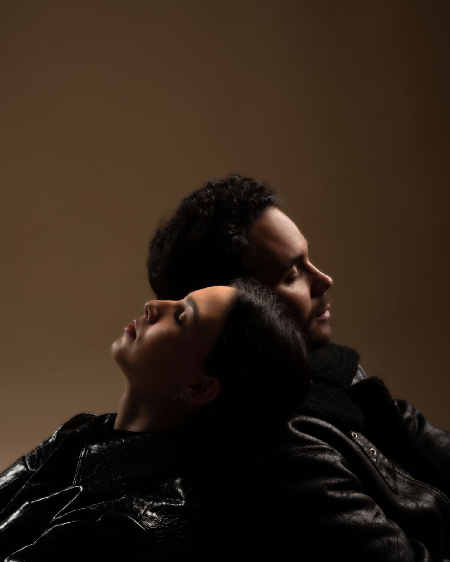 Romantic couple in black leather jackets posed back-to-back looking upward in dramatic moody lighting.