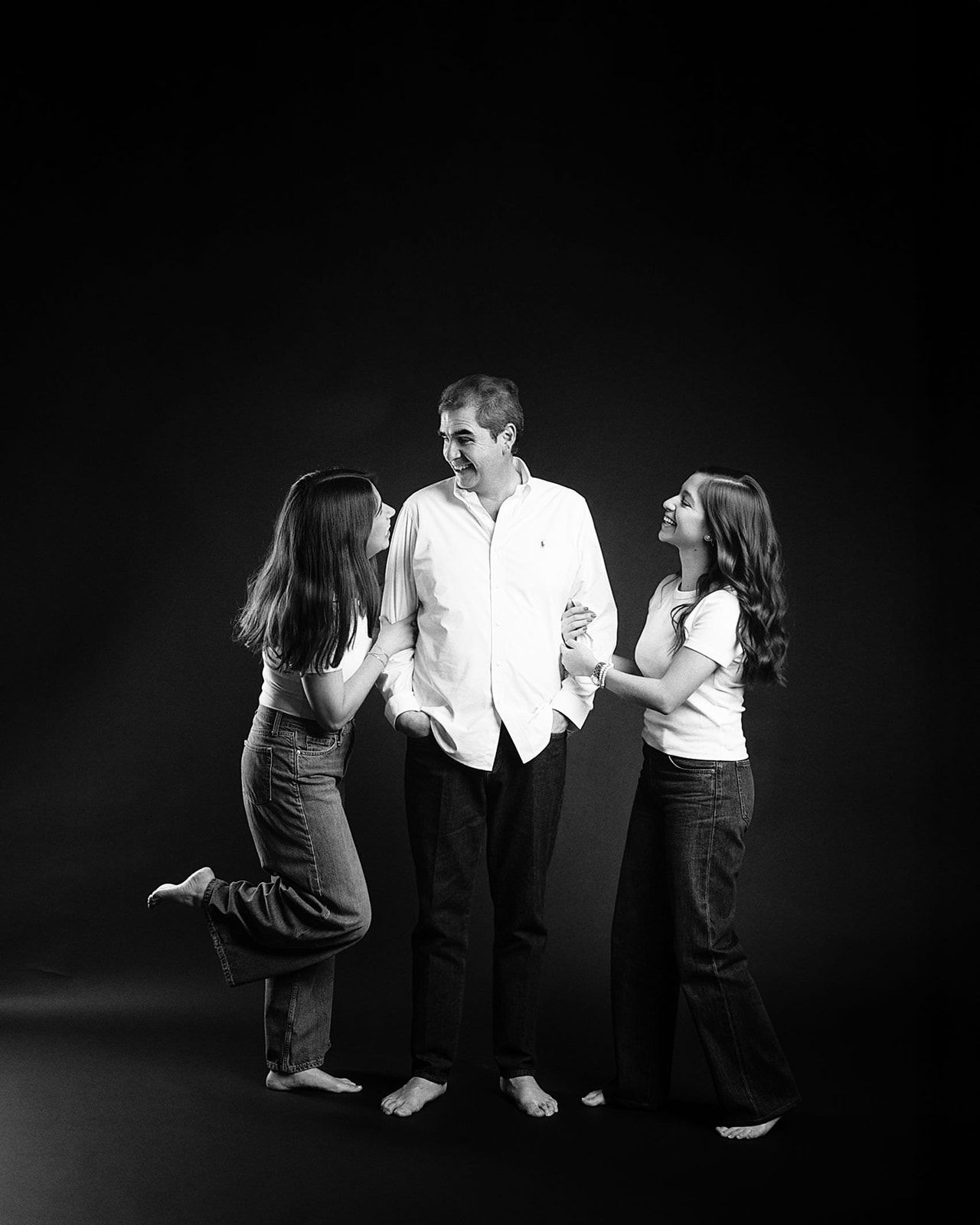 Black and white studio photo of a man standing between two young women in casual white tops and jeans.