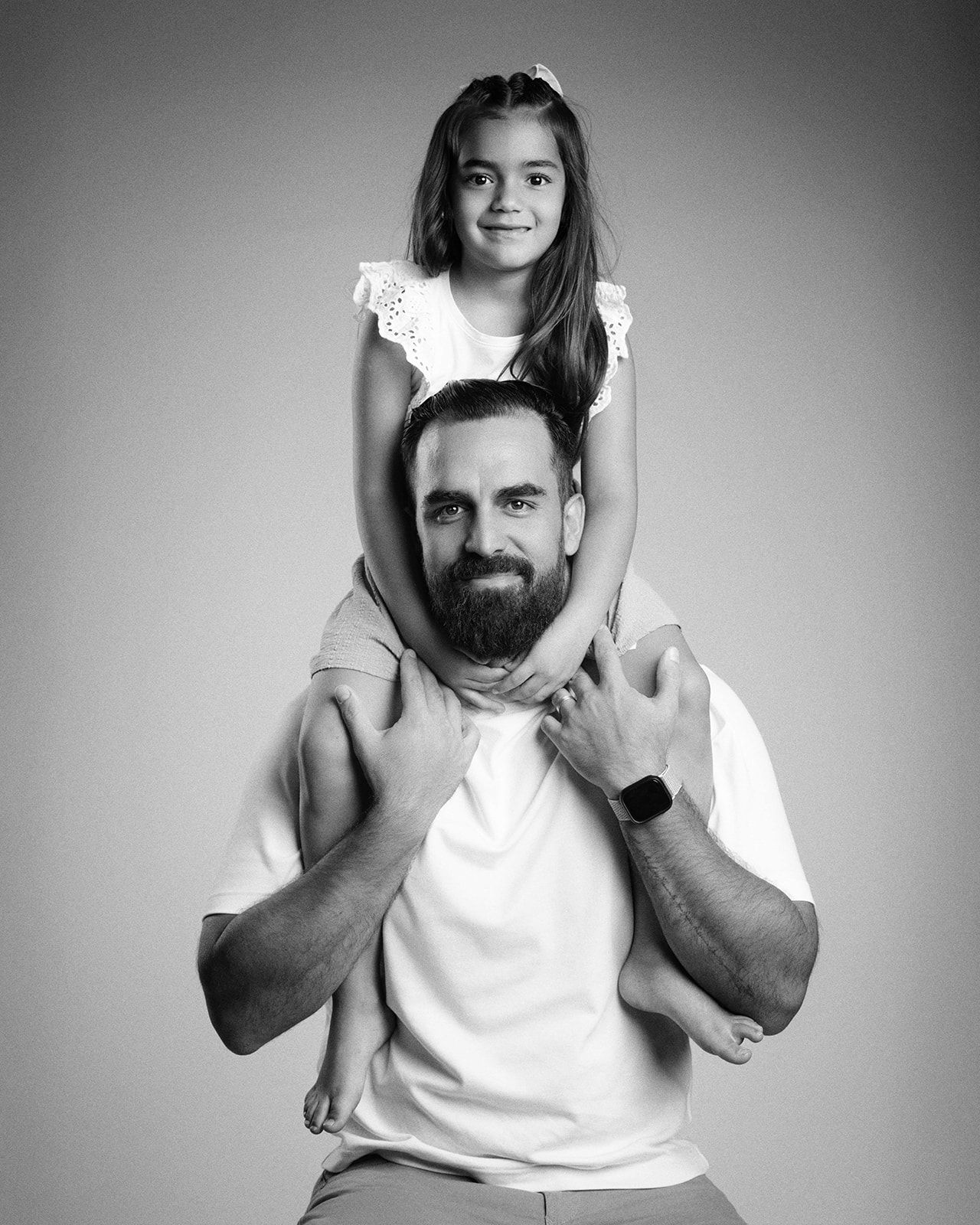 Black and white portrait of a bearded father carrying a smiling young girl on his shoulders in a studio.