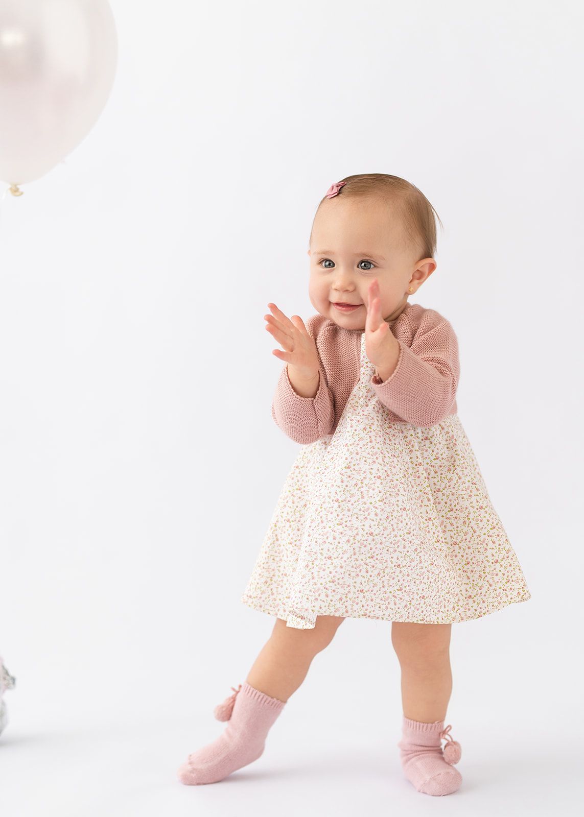 Happy baby girl in pink cardigan and floral dress clapping hands next to a white balloon.