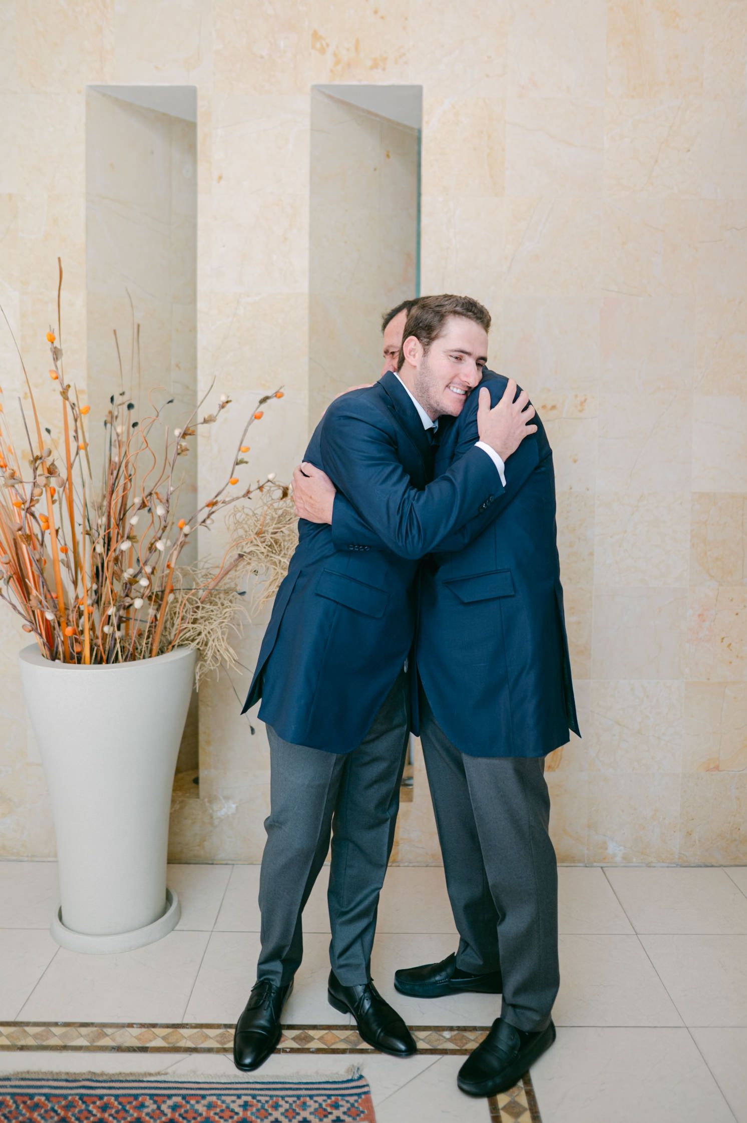 Two groomsmen in navy suits sharing an emotional hug before the wedding ceremony.