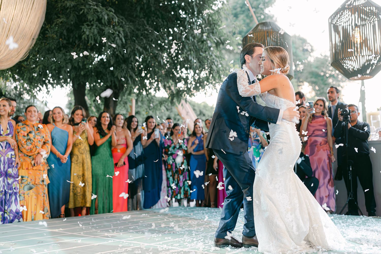 Bride and groom kiss as guests toss white confetti petals during a joyful outdoor wedding celebration.