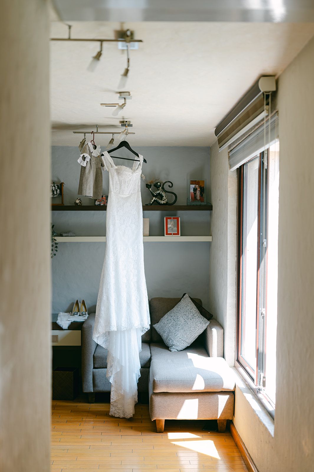 White lace wedding dress hanging in sunlit room with bridal shoes below.