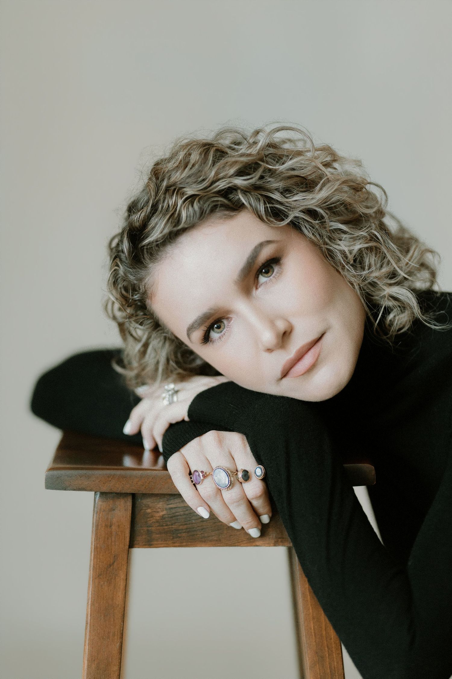 Close-up of curly-haired woman in black turtleneck resting on stool, wearing colorful rings.