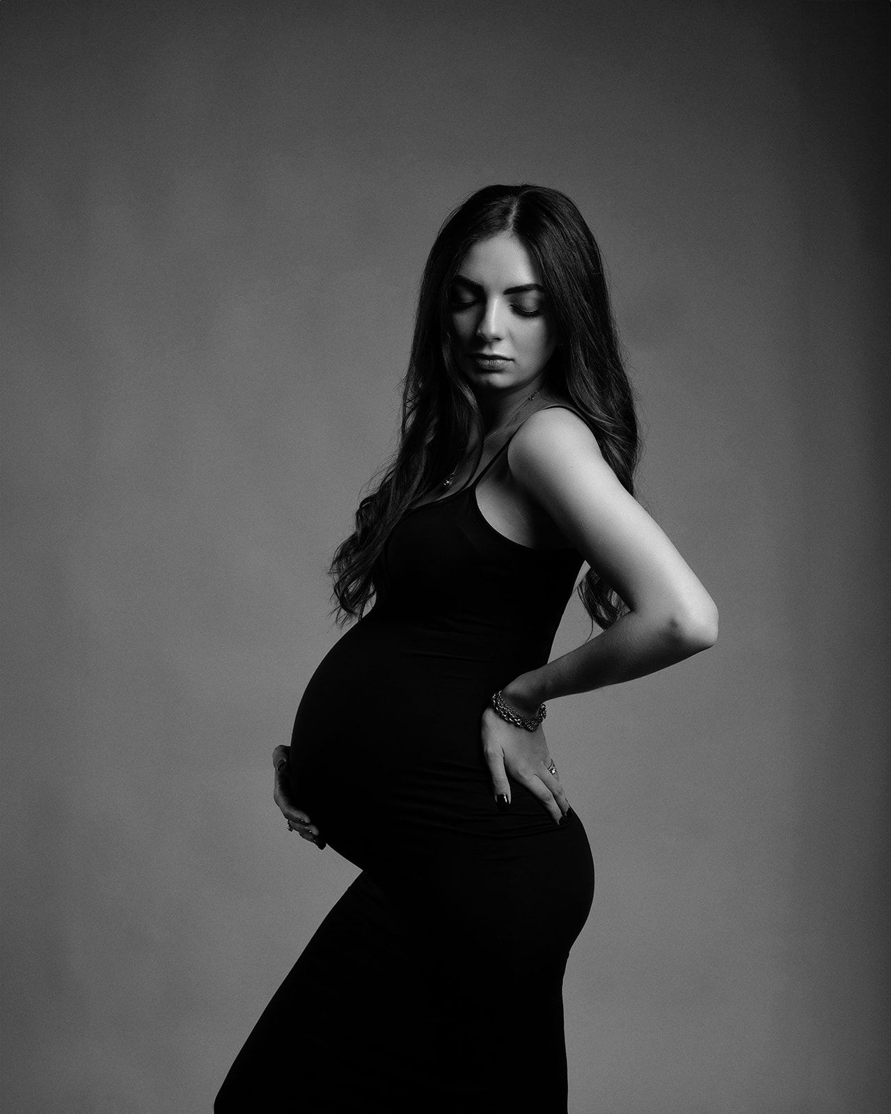 Elegant black and white maternity portrait of a pregnant woman in a black dress with eyes closed.