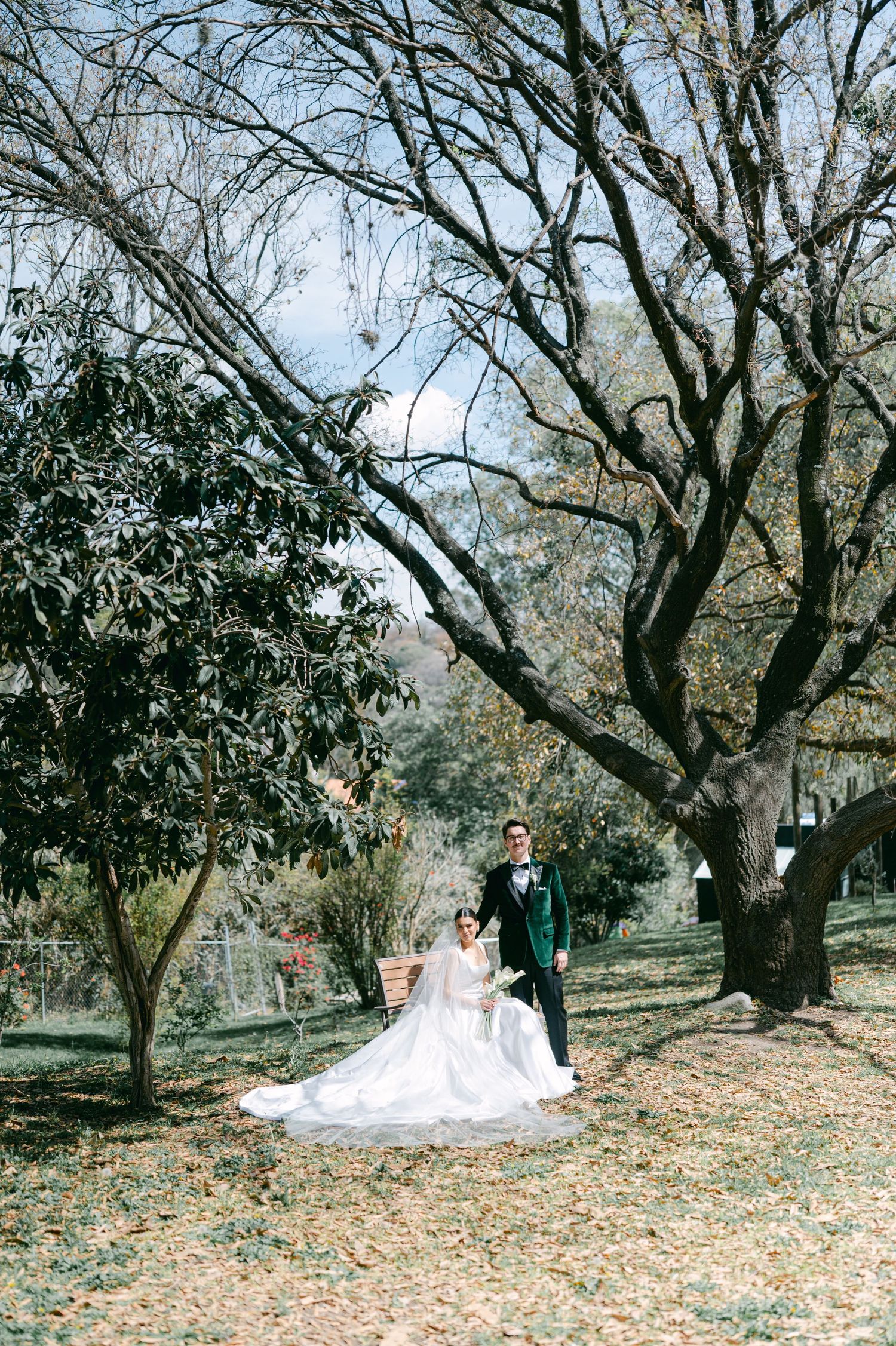 Wedding couple walks hand in hand through autumn garden, bride in flowing gown with long veil, groom in green velvet suit.