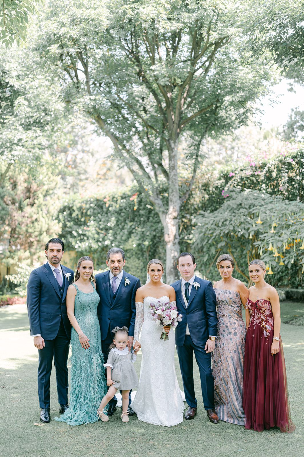 Wedding party group portrait outdoors featuring bride, groom, and family in formal attire on green lawn.