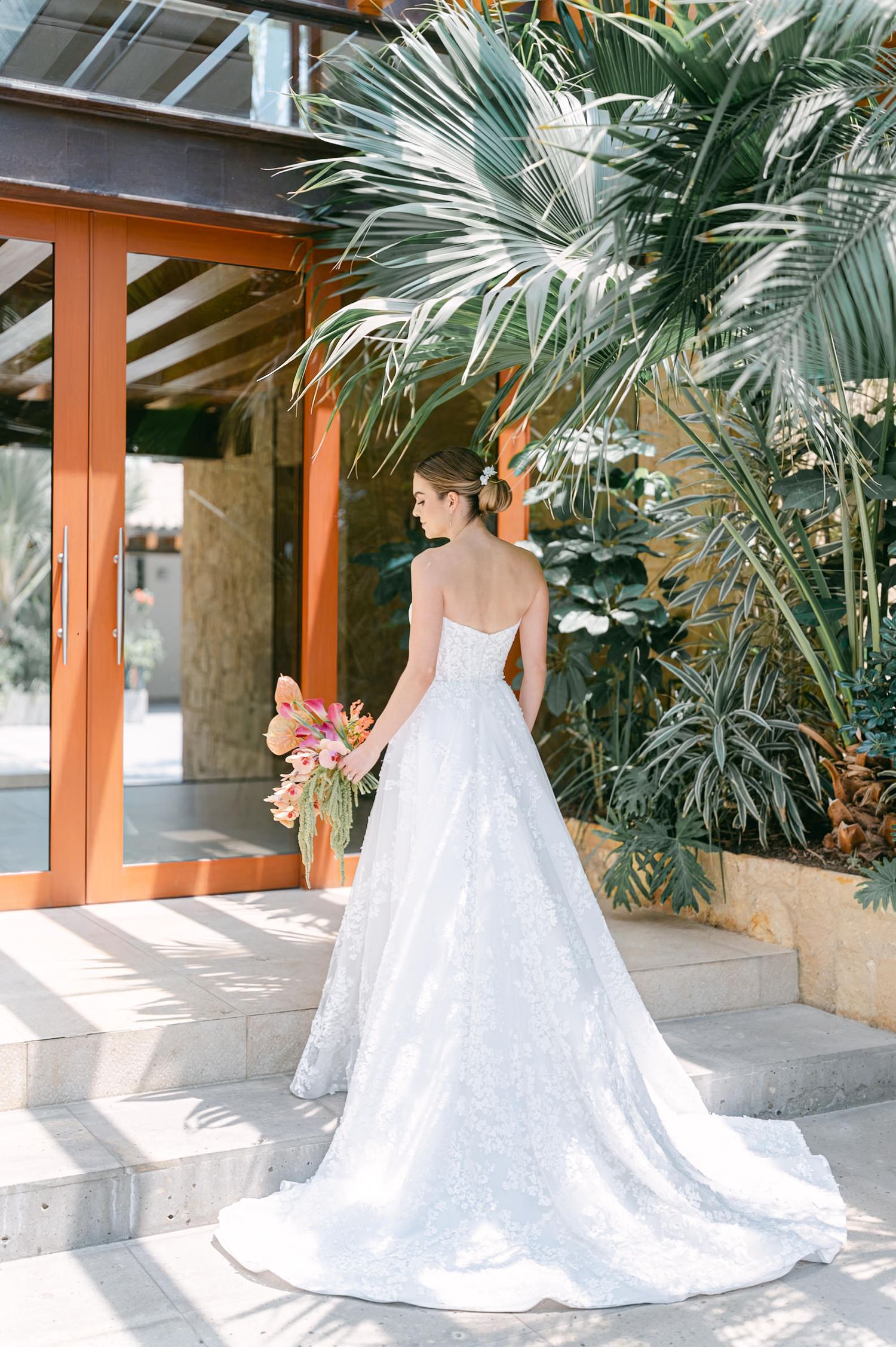 Bride in strapless lace gown with long train standing on steps surrounded by tropical palm leaves.