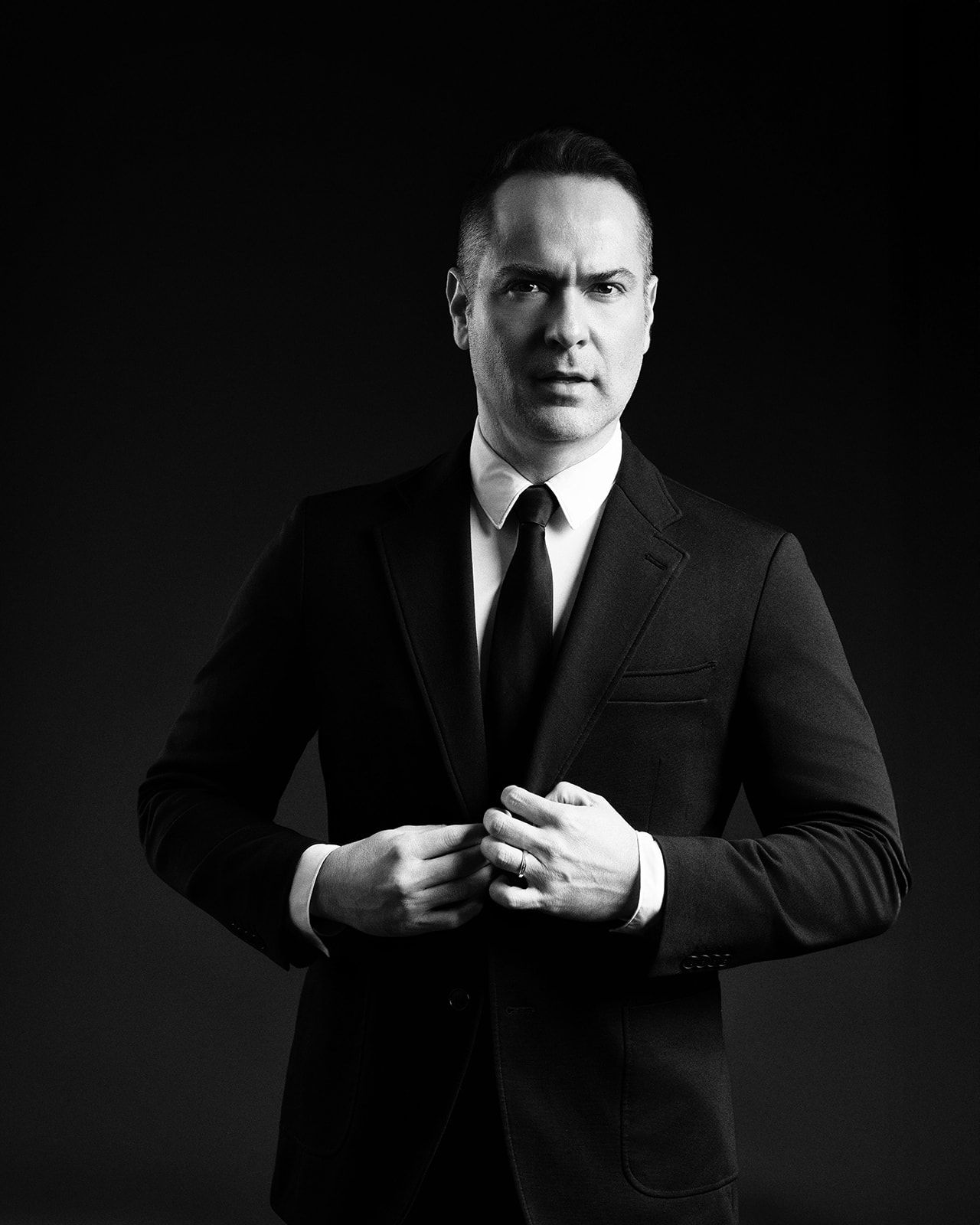 Black and white portrait of a serious man in a black suit adjusting his jacket lapels.