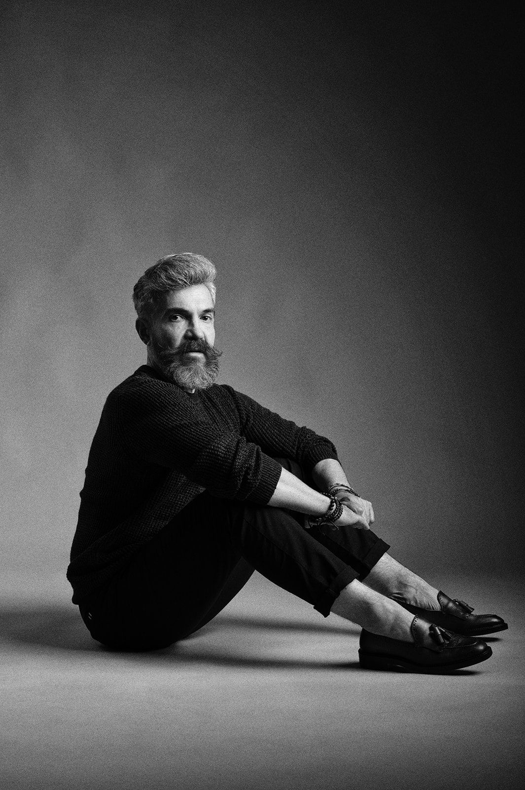 Black and white portrait of a bearded man in all-black outfit sitting on floor, looking at camera.