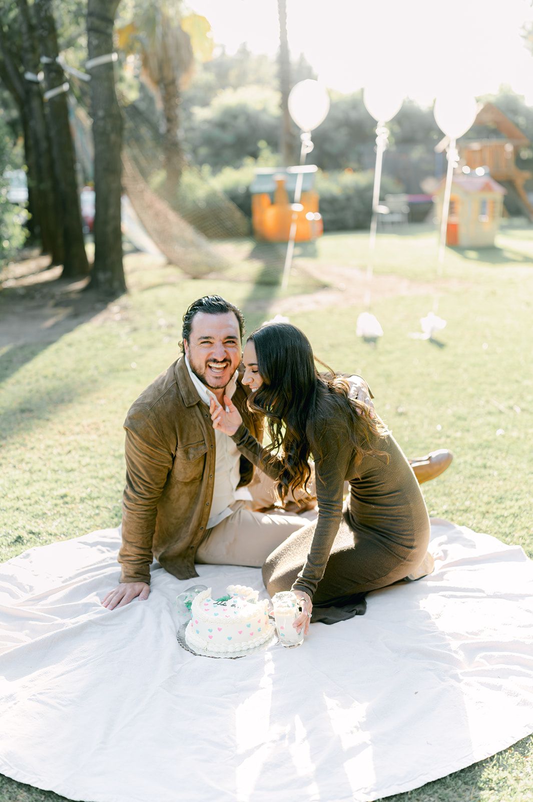 Couple laughing together on picnic blanket with gender reveal cake, woman kissing man on cheek outdoors.