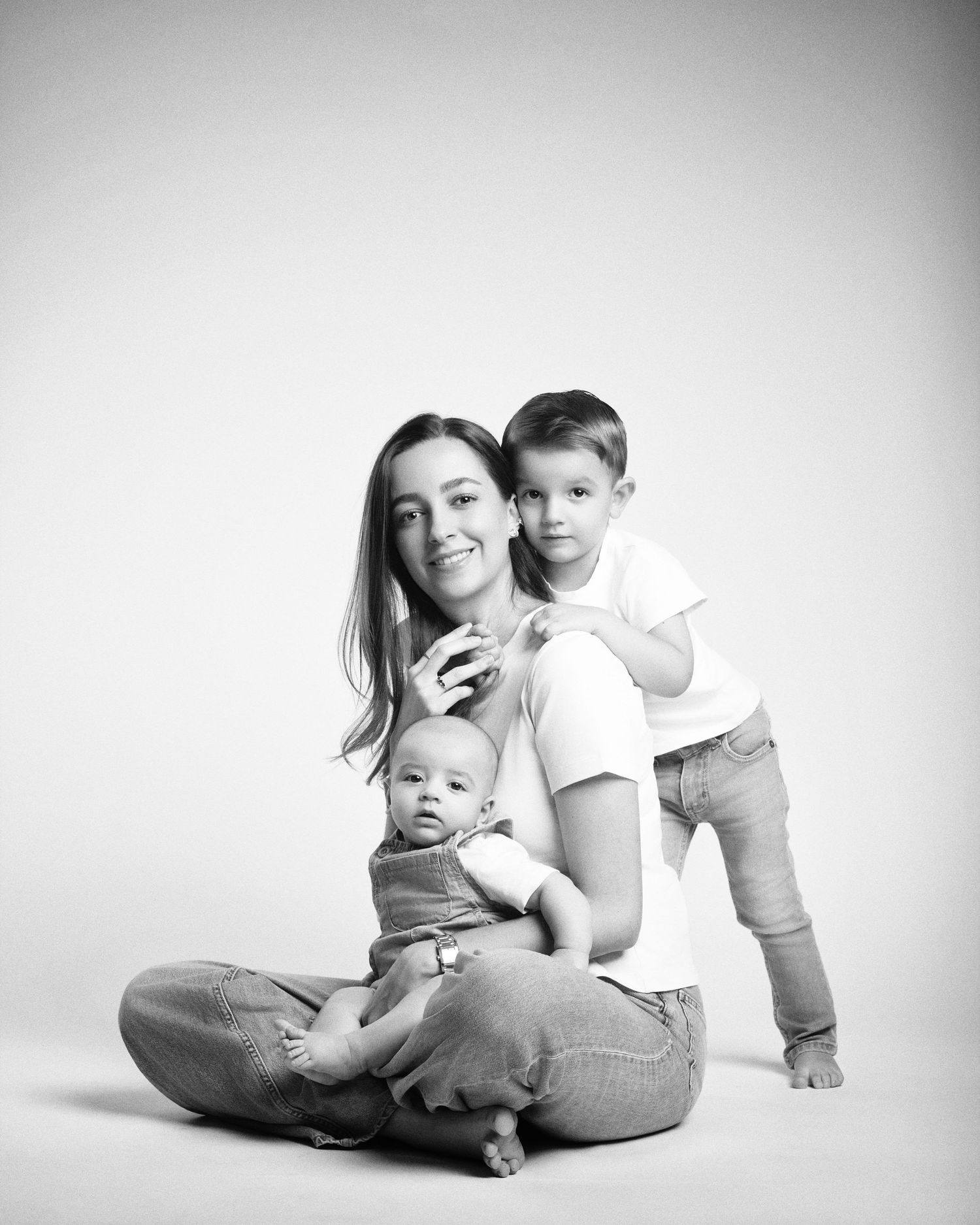 Black and white studio portrait of a mother sitting with her baby and toddler son.