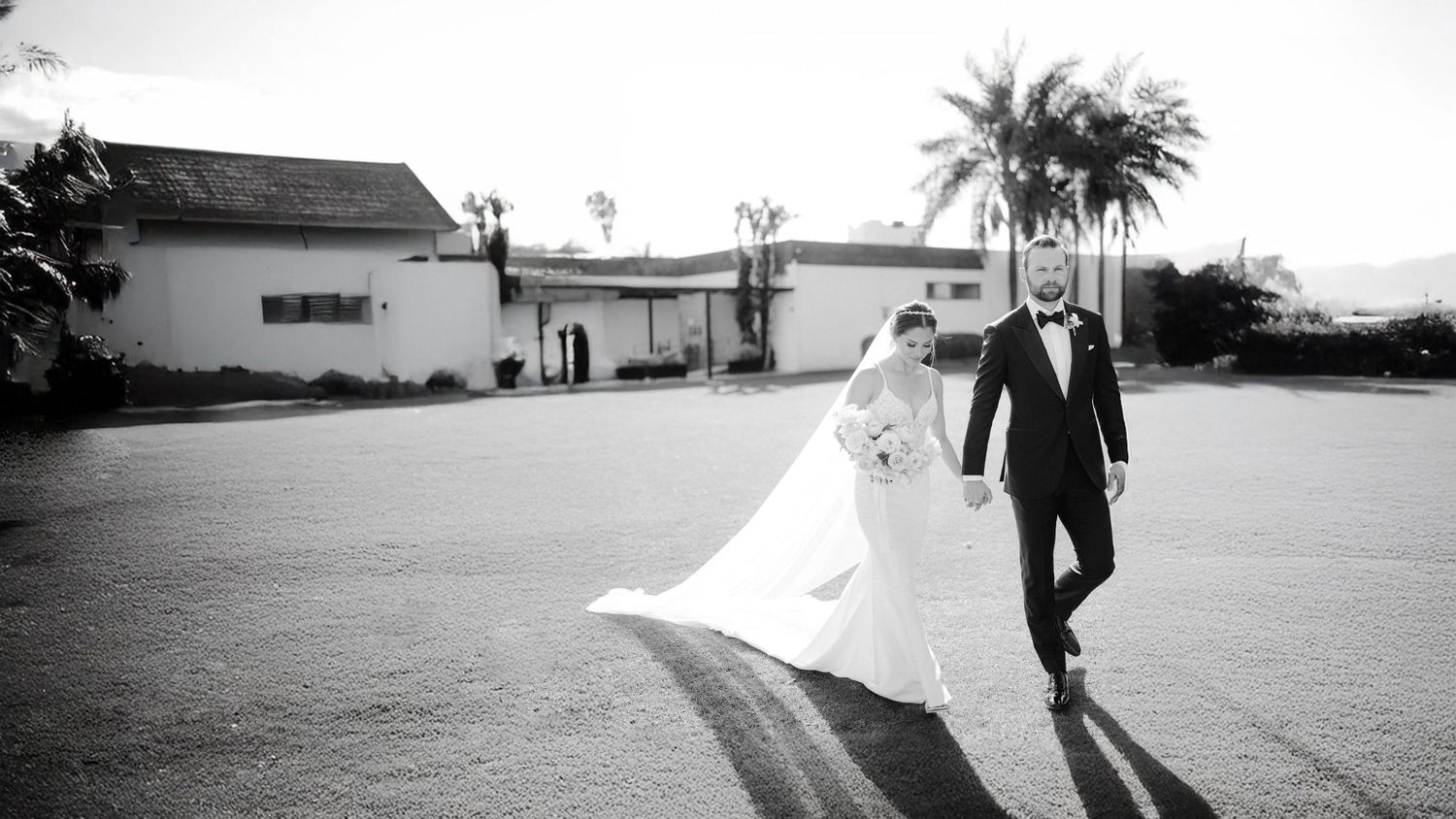 Black and white photo of a bride and groom walking hand in hand outside a building on their wedding day.