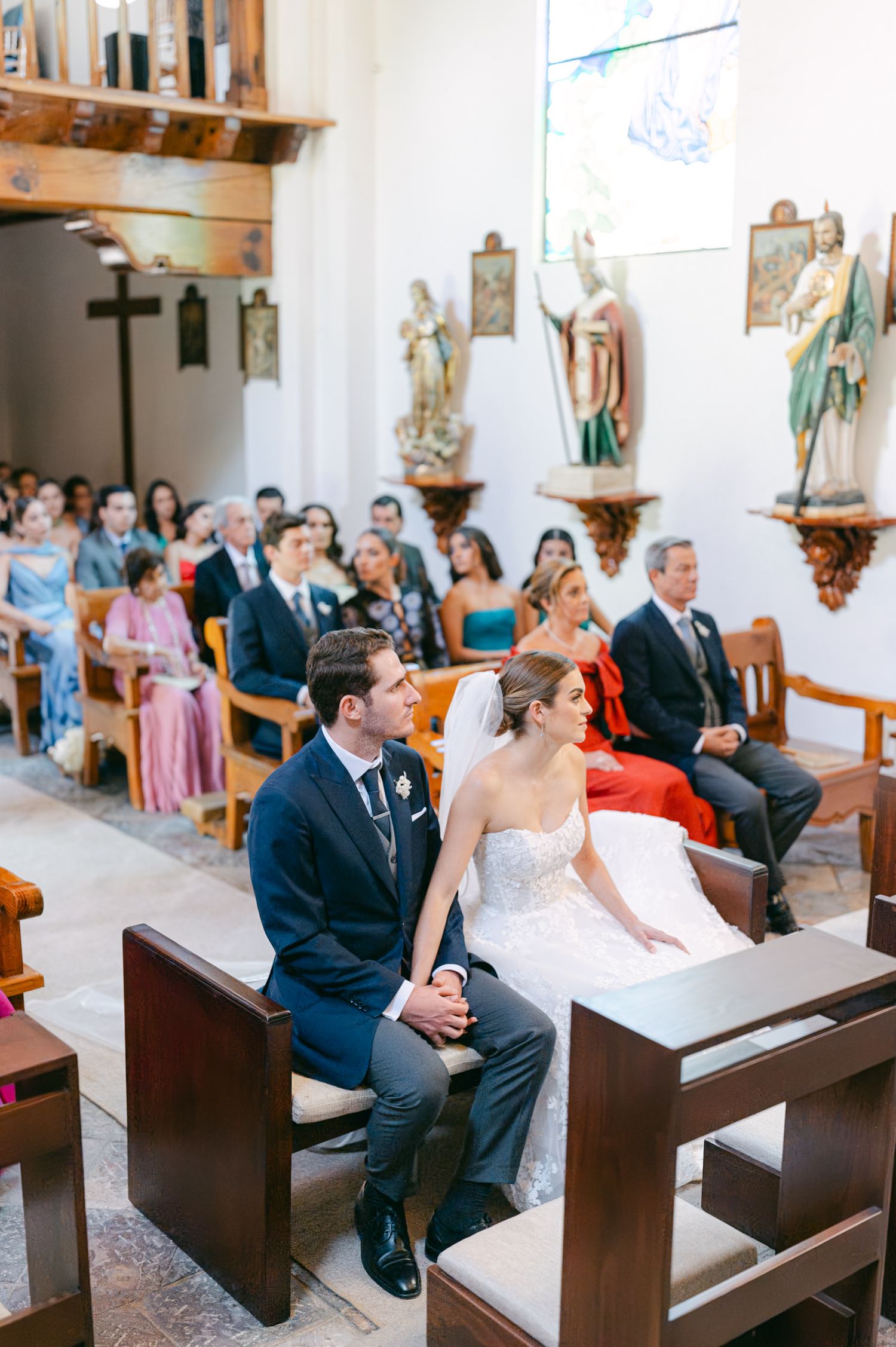 Wedding ceremony in a traditional white chapel with wooden pews and religious artwork on walls.