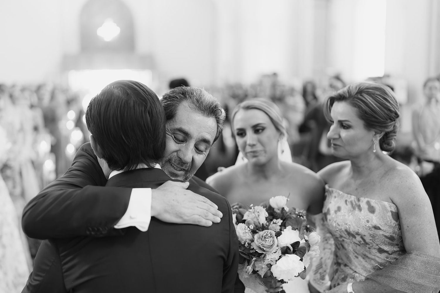 Black and white photo of groom embracing a guest as bride holds bouquet at church ceremony.