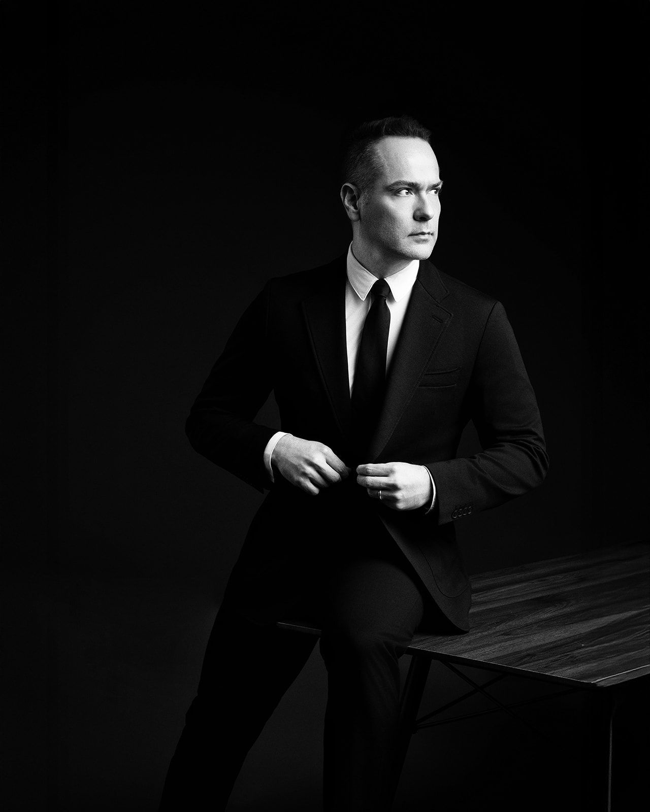 Elegant man in a black suit and tie posing in a dramatic dark studio setting.