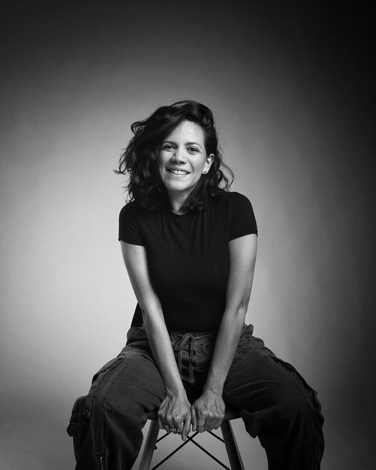 Black and white portrait of a laughing dark-haired woman seated in a black t-shirt and cargo pants.