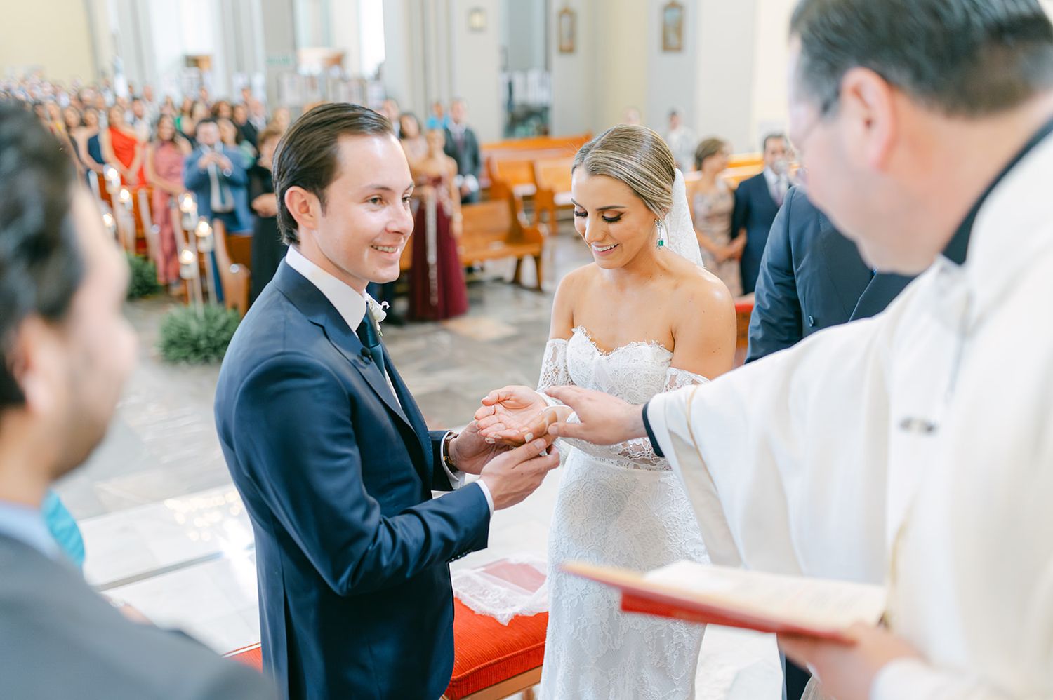 Smiling groom receives wedding ring from priest as bride watches during church ceremony.