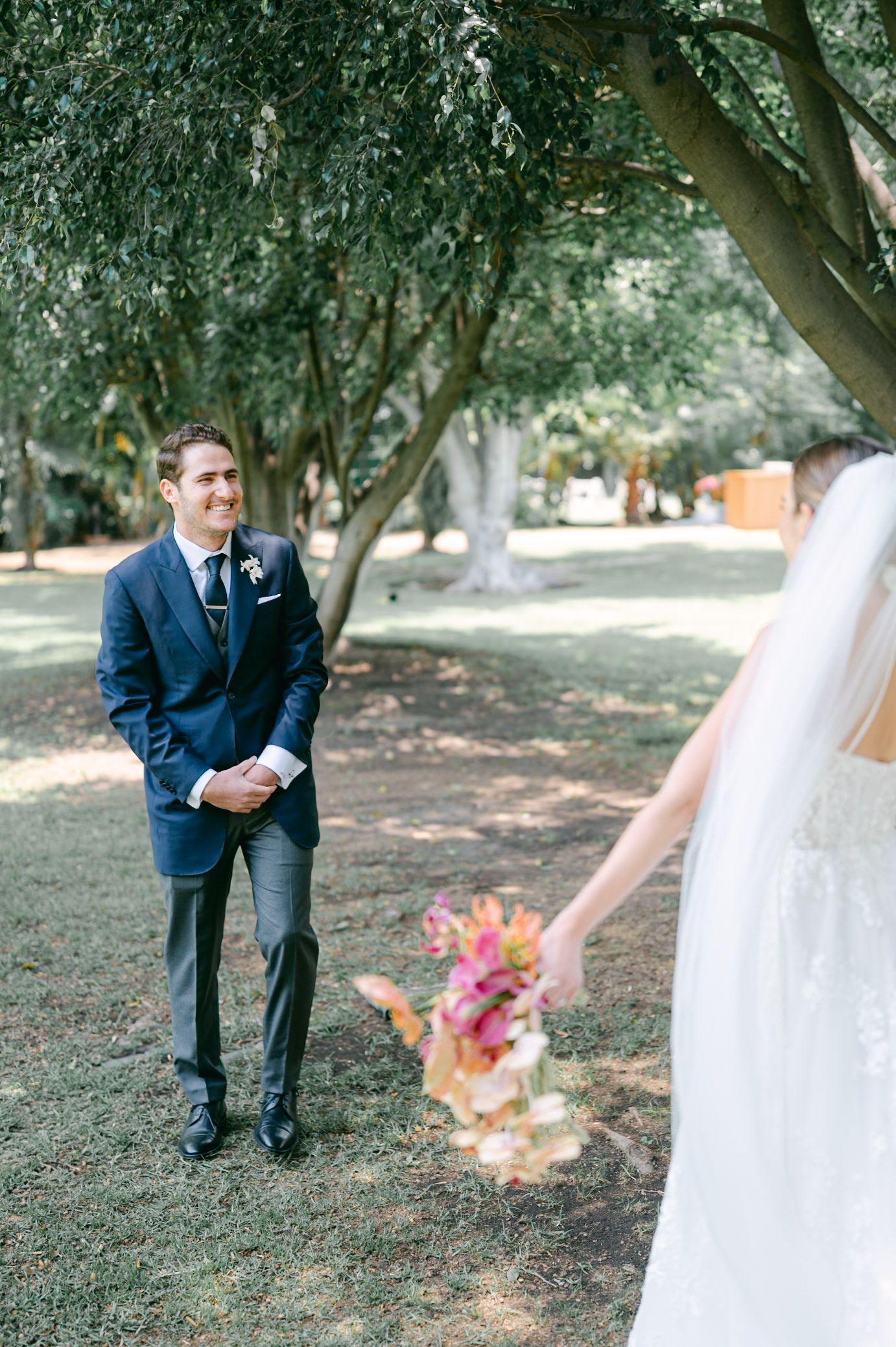 Groom in navy suit smiling at bride during first look moment under shaded trees outdoors.