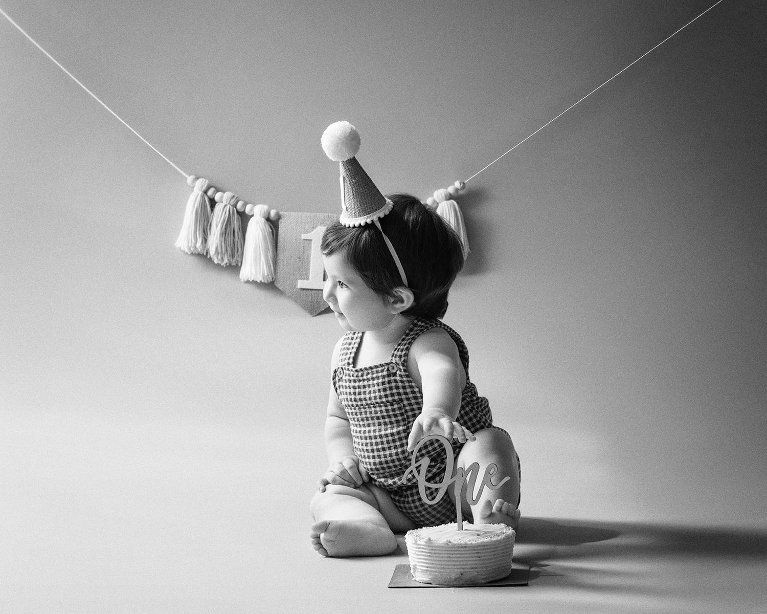 Black and white photo of a baby in a party hat celebrating their first birthday with a smashing cake.