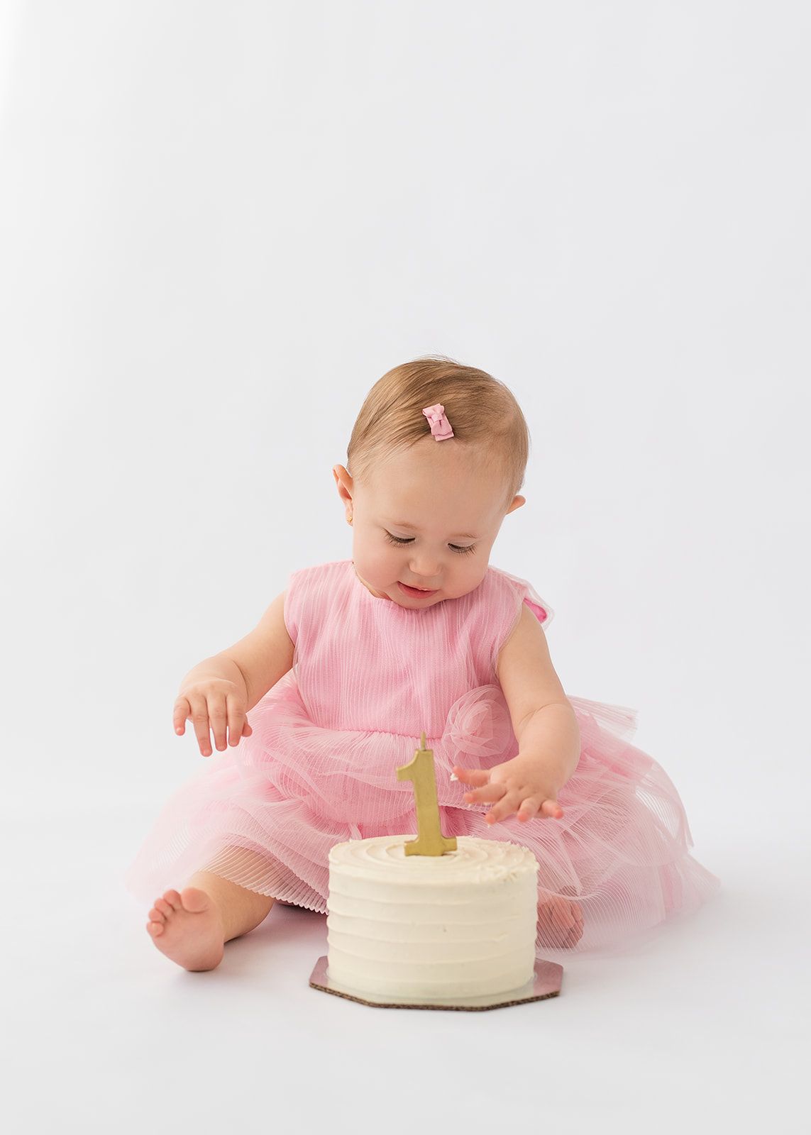 Baby girl in pink tulle dress explores her first birthday smash cake with gold number one candle.