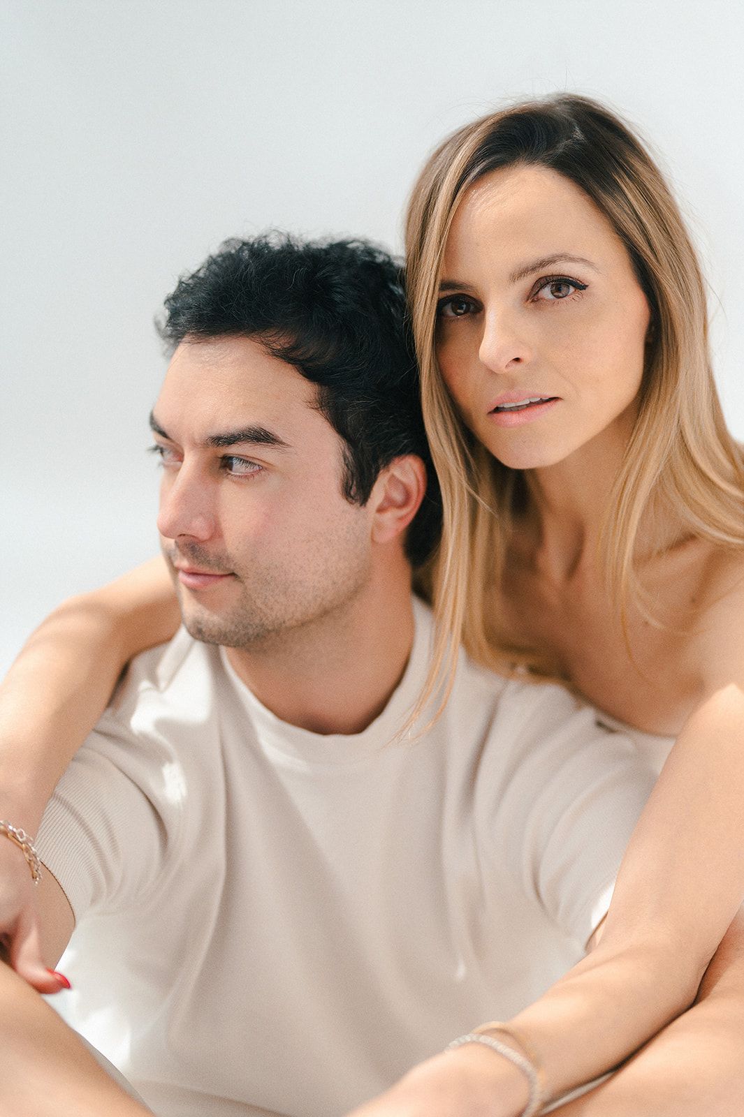 A man and woman pose together against a white background, she rests her chin on his shoulder looking at camera.