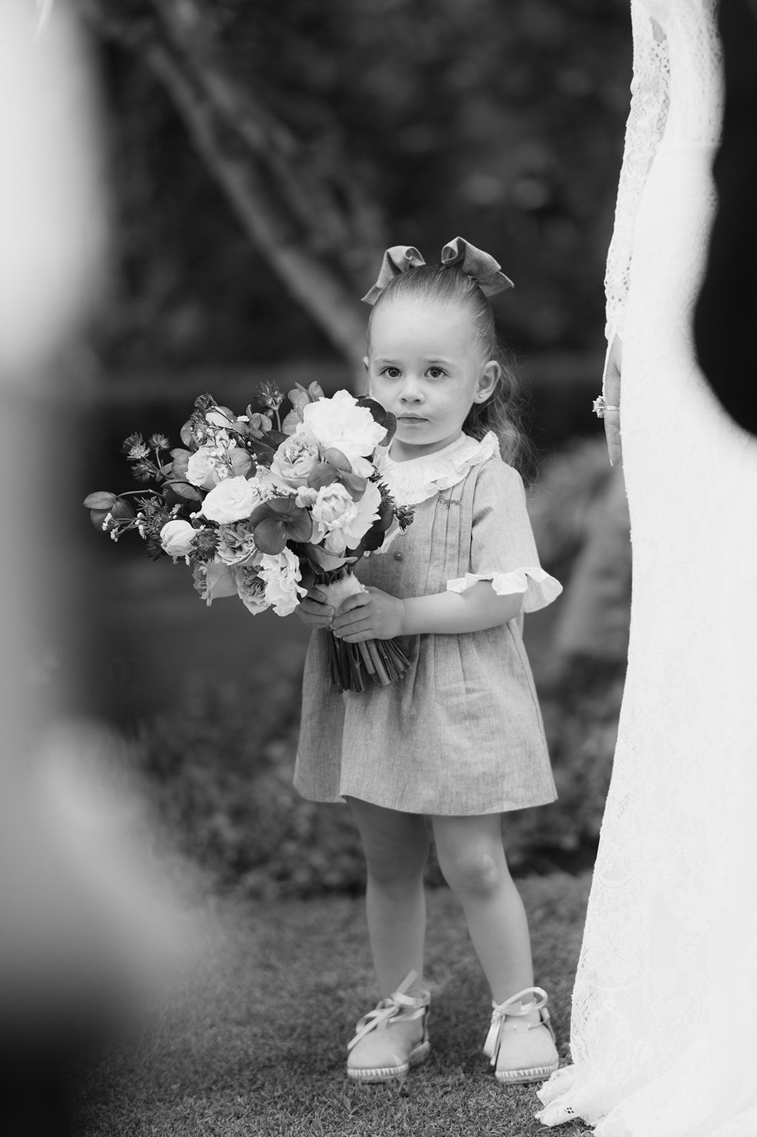 Adorable flower girl in linen dress holds floral bouquet at outdoor wedding ceremony.