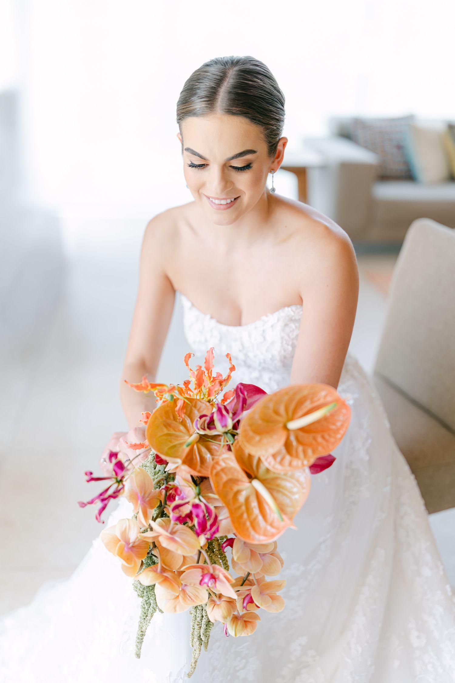 Radiant bride in strapless lace gown smiling down at vibrant tropical orange and pink bouquet.